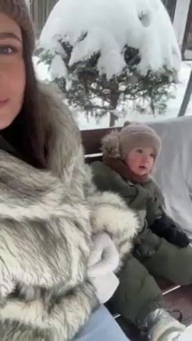 The video shows a woman and a small child sitting on a wooden bench in a snowy outdoor environment. The woman, on the left, is wearing a brown knitted beanie, a grey and white faux fur coat, and white gloves. She is looking at the camera and smiling slightly. To her right, the child is wearing a brown knitted beanie with a faux fur pom-pom, a brown scarf, and an olive green snowsuit with a fur-lined hood. The child appears to be looking off to the right side of the frame.  Behind them, a tree is heavily laden with snow. The ground is also covered in snow.  The camera is mostly still, with slight movements, suggesting a selfie or self-recording perspective. The lighting is soft and diffused, typical of a snowy day.