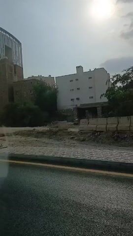 The camera, likely from a moving vehicle, captures a street view under a bright, overcast sky. Buildings of varying sizes and architectural styles line the street. The foreground shows the road with a painted yellow line marking the edge. Adjacent to the road is a paved sidewalk. Beyond the sidewalk, there is an unpaved area with construction debris. A light post stands between the sidewalk and the unpaved area. In the distance, buildings are visible. The video progresses as the camera moves, changing the perspective on the scene. The sun is visible through the clouds. The buildings are primarily white or beige and of medium height. The terrain beyond the road and sidewalk shifts from flat, unpaved surfaces to a slope reinforced with concrete and metal elements. The street appears to be in a state of transition or redevelopment.