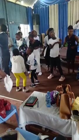 The video shows a group of people dancing in an indoor space with a wooden floor and a ceiling covered with light blue and white fabric. A table covered with a white tablecloth is visible in the foreground, with several items on it including a water bottle, books, and bags. The people, including children and adults, are dressed in casual attire, and some are wearing white t-shirts. They appear to be engaged in a lively dance. The overall atmosphere seems celebratory.
