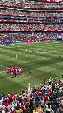The video shows a crowded stadium during a soccer match. Spectators fill the tiered seating, many wearing red and white. On the green field, a group of players in red uniforms are huddled together in the center, appearing to celebrate or strategize. Other players in yellow and green uniforms are spread across the field, and a few individuals in suits are observing. A cameraman is positioned near the huddle, filming the scene. The environment appears to be outdoors, with bright sunlight illuminating the stadium.