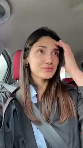Here is a concise description of the video.
The video shows a woman in a car. She has long brown hair and is wearing a black jacket and a light blue shirt. She is wearing a seatbelt and smiling. She is running her hand through her hair. The interior of the car is visible, including the red headrests and gray ceiling.