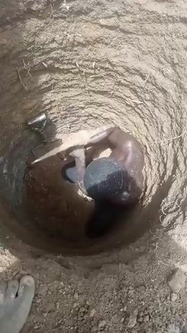 The media file shows a person in a pit, seemingly digging. The scene is set in a dusty, outdoor environment, likely DU, Nigeria, given the location context. The pit is circular and dug into the earth, with visible soil and some sparse vegetation. A small metal tool, possibly a shovel or trowel, rests at the top edge of the pit.

A person, whose skin appears wet and shiny, is partially submerged in the pit. They are crouched down and their arms are engaged in activity within the pit. The lighting suggests it is daytime, but the specific time of day and weather conditions are not discernible.

The person's body is mostly visible, though their head is obscured by shadow or their position. They are wearing dark shorts. Another dark, roughly spherical object is next to them in the pit. The overall impression is one of manual labor in a confined, earthen space. There is no visible text in the media file.