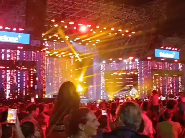 The video shows a large crowd of people at an outdoor concert or festival. The stage is illuminated with bright lights, including red and yellow spotlights, and a large screen displaying abstract patterns. There are also smaller screens displaying the "ticketone" logo. People in the crowd are holding up their phones to record the event. The overall atmosphere appears to be energetic and celebratory.