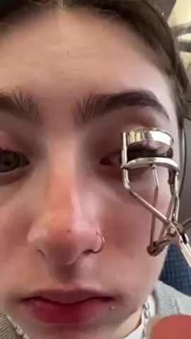 A close-up shot shows a fair-skinned young woman with dark hair pulled back, wearing headphones, and using an eyelash curler on her left eye. She has a nose ring in her left nostril and freckles across her nose. The background suggests she is on a train or bus, with a patterned seat visible. The focus is primarily on her face and the eyelash curler.