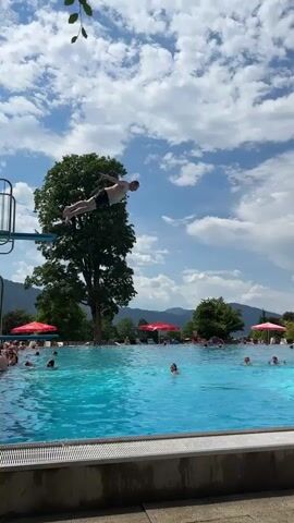 A person is captured mid-air, performing a dive into a bright blue swimming pool. They are positioned horizontally, with arms extended, and appear to be wearing dark shorts. The dive is happening on a sunny day, with fluffy white clouds dotting a blue sky. A large, leafy green tree stands tall to the left of the frame, behind a blue diving board. In the background, a group of people are visible in the pool and around its edges, some relaxing under red umbrellas. Further in the distance, rolling green hills and mountains are visible under the expansive sky, creating a scenic outdoor setting. The camera angle is from below the pool's edge, looking upwards towards the diver and the sky.