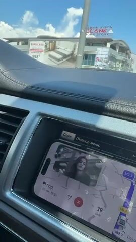 The video shows the interior of a car as it drives on a sunny day. The driver, a man wearing sunglasses, is holding the steering wheel. The dashboard displays a navigation map on a phone. Outside the car, buildings and other vehicles are visible. The camera pans slightly, revealing a billboard advertising a home store.