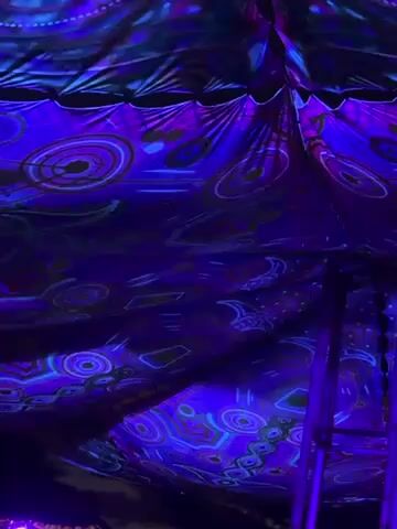 The video shows the interior of a tent or canopy that is decorated with a swirling psychedelic pattern and illuminated with bright purple and blue light.  Green laser beams shine upward, reaching outside the tent and into the night sky. The sky is dark, and a few clouds are visible.  The video appears to be taken at a music festival or party.