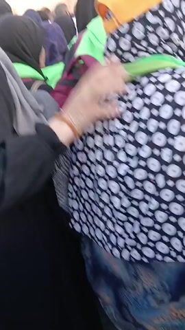 The video shows a crowd of people gathered in front of the Kaaba in Mecca. The Kaaba is a large, black, cube-shaped building adorned with gold. Many of the people in the crowd are wearing Muslim head coverings, including hijabs and headscarves. Some of the men have shaved heads. Several people are wearing bright green vests. Some people are holding books. The video appears to be taken from within the crowd, giving the viewer a close-up perspective of the people and the Kaaba.