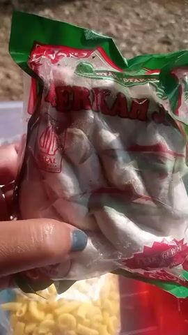 A hand with grey nail polish holds a clear plastic bag of white, oval-shaped food items. The bag has a green and red striped top and features text and logos, including a red "HALAL" symbol. Below the bag of food, a portion of a clear plastic container filled with yellow, macaroni-shaped pasta is visible. The background appears to be outdoors, with blurred green and brown elements suggesting foliage and possibly a surface.