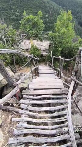 A first-person perspective video shows a person walking down a narrow, rustic wooden staircase that clings to the side of a rocky, forested mountain. The staircase is made of weathered planks and has rough-hewn wooden railings. The surrounding environment is lush green with trees and vegetation growing on the steep slopes. The sky appears overcast with clouds. The person is wearing athletic shorts and white sneakers.