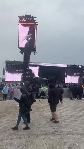 An outdoor event space under an overcast sky features a large stage setup with multiple towering video screens in the background. The screens display graphics with pink and purple hues, and text such as "Spiritbox," "Hamburg," and "Sportplatz" along with a "T-" logo. The ground is paved with light-colored square tiles. Numerous individuals, mostly adults, are scattered across the paved area, many wearing dark jackets, coats, and hoodies. They are seen standing, walking, and milling about. Barriers or fencing are present in the middle ground. A person in a black hooded jacket is in the foreground, facing away from the camera.