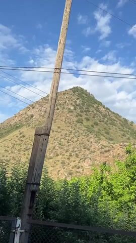 The video captures a view of a mountain landscape on a sunny day. The foreground is dominated by a wooden utility pole and lush green trees. In the midground, a brown, rocky mountain slope is visible, sparsely covered with vegetation. The sky above is a vibrant blue, dotted with scattered white clouds. In the distance, more rugged hills and rock formations are seen. Power lines are strung across the upper portion of the frame, crossing over the mountain and trees. The scene appears to be in a rural or natural setting.