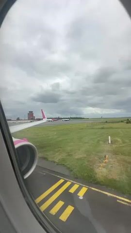 The video, filmed from an airplane window, captures a ground-level view of an airport on an overcast day. In the immediate foreground, the window frame encircles a portion of a pink-and-white airplane wing and its engine, suggesting the aircraft is likely a Wizz Air plane. The plane appears to be stationary or slowly taxiing on a tarmac section, marked with distinct yellow lines.

Beyond the wing, the airport environment unfolds. A grassy expanse separates the viewer's aircraft from other parts of the airfield. Several other aircraft are visible in the distance: at least one more with a pink tail fin, and another white and blue plane, all parked or moving slowly on distant taxiways. A distinctive reddish-brown control tower stands prominent on the left side of the frame, hinting at the airport's specific architecture. Given the context, this is likely Beauvais–Tillé Airport in Tillé, France. The sky is entirely covered with thick grey clouds, indicating overcast weather during daylight hours. No people are visibly depicted. Muffled, indistinct human speech can be heard, likely from inside the cabin.