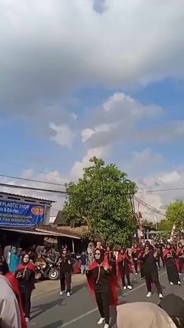A group of people march in a parade on a sunny day with scattered clouds. They are dressed in black outfits with red capes and black headscarves. They appear to be performing a choreographed dance. Spectators line the sides of the road, watching the parade. A plastic shop is visible in the background. The overall atmosphere seems festive and celebratory.