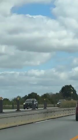 The video captures a view from a moving vehicle on a highway. The sky is filled with fluffy white clouds against a bright blue backdrop. On the right side of the road, a red SUV is in focus, its rear is visible. Further down the road, a black SUV is seen driving in the opposite direction. To the left, the highway is divided by a concrete barrier with bollards. Beyond the barrier, a line of trees and vegetation borders the road. A solar-powered traffic sign with a yellow circular indicator is also visible in the distance.