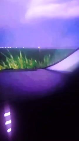 The video shows a view from inside a vehicle looking out at a blurry field of green vegetation under a purple sky. The foreground is dark and out of focus, with some glowing purple lights visible on the left side. In the distance, there are faint white lights scattered across the horizon, possibly indicating a town or other structures. The overall scene is somewhat abstract due to the blurriness and the purple lighting.