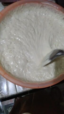 The video shows a close-up view of a person stirring a large bowl of what appears to be a dough or batter. The bowl is an orange plastic basin. The stirring is being done with a metal spoon, and the mixture is foamy and has small bubbles throughout. The motion of the spoon creates swirls and peaks in the mixture. The background is out of focus but suggests an indoor setting with some patterned surfaces and what might be wooden furniture. The lighting suggests it is likely daytime. The video appears to be recorded in Rambipuji, Indonesia.
