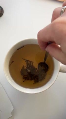 A hand with a silver ring stirs a mug of brown liquid with a spoon. The mug is white and contains tea leaves. The table is white. There are a few sheets of paper and a metallic object nearby.