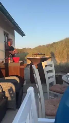 A man is grilling outside next to a house. He is standing behind a wooden table with a red bowl and spices on it. The grill is round and contains some food. There are several chairs and a table set for dinner nearby. Behind the grill is a grassy dune. The sky is clear and blue.