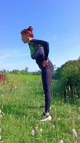 A woman with red hair pulled into a bun is performing back extensions outdoors. She is wearing a black long-sleeved athletic top with green accents and black leggings. Her hands are on her hips. She is standing in a grassy field with some plants and a blue sky in the background. The video is a compilation of short clips showing her bending backward and then returning to an upright position, repeating the motion.