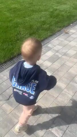 The video shows a young child walking away from the camera on a paved walkway. The child is wearing a dark blue hoodie with writing on the back and light pink pants. The walkway is made of light-colored rectangular pavers and has a drain grate along the edge. To the left of the walkway is a patch of green grass. In the background, there is a fence and some stacked wood. The sun is shining, casting shadows on the ground. The child appears to be walking towards a gate or an open area.