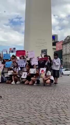 A group of young people are kneeling and squatting in a plaza, holding up signs and pictures in front of them. They are facing forward, and one person to the side seems to be reading from a paper. In the background, there is a tall, white obelisk-like structure with inscriptions on it. The plaza is paved with brickwork in a herringbone pattern. Above them, the sky is partly cloudy. A bird walks in the foreground, seemingly unfazed by the group.