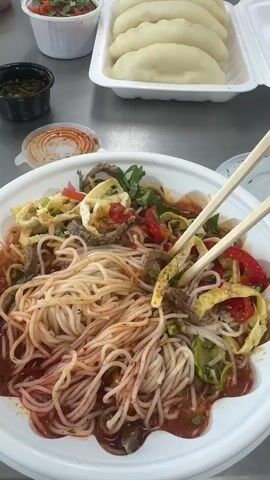 The video shows a close-up of a bowl of noodles with chopsticks in it, as well as other food items on a metal table. In the background, several people are seated at the table, eating. The environment appears to be a communal dining area or a cafeteria. The lighting is bright, suggesting an indoor setting during the day. The overall atmosphere seems casual, with people enjoying a meal together.