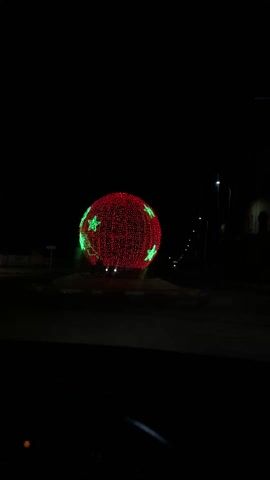 The video depicts a large, spherical decorative object at night, likely a holiday display. The sphere is primarily illuminated with red lights and features green star shapes distributed across its surface. It is positioned in the center of a traffic roundabout. The surrounding environment is dark, with a few streetlights visible in the background, along with some buildings and signage. The video appears to be recorded from inside a vehicle, based on the perspective and the dashboard visible at the bottom of the frame.