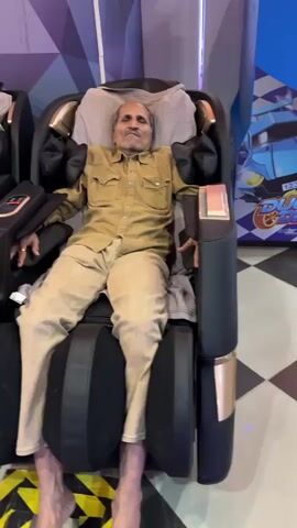 An elderly man is sitting in a black and gold massage chair. He is wearing a beige button-down shirt and beige pants. The chair has what appear to be air bladders on the sides that are pushing against his arms and legs. The man has his eyes closed and appears to be either asleep or trying to relax. The chair is located in an arcade, with other massage chairs visible nearby, as well as an arcade game. The floor has black and white checkerboard tiling. There is a yellow striped caution tape around the chair. Loud arcade sounds can be heard in the background.