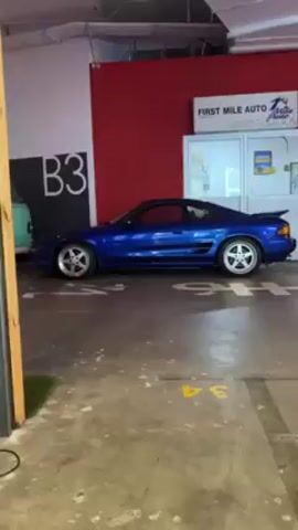 The video shows a parking garage with a red wall and a garage door. Two blue cars are parked in the garage, one is a Toyota MR2 and the other is a BMW E36. There is a sign for "First Mile Auto". The garage has yellow painted parking space numbers. The camera moves from left to right to show the cars.