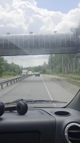 A POV shot from inside a vehicle shows a road with traffic. The sky is cloudy with patches of blue. Trees line the sides of the road, with some birch trees visible on the right. A pedestrian overpass is visible above the vehicle. Two black headphones are resting on the dashboard in the foreground. Cars and trucks are driving on the road.