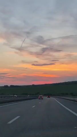 The media file shows a view from a car driving on a highway at sunset. The sky is filled with colorful clouds, ranging from pale blue and gray to vibrant orange and pink. There are several contrails visible in the sky, suggesting the presence of airplanes. The sun is low on the horizon, casting a warm glow over the landscape.

The scene depicts a highway with multiple lanes and a metal guardrail on the side. A few cars are visible on the road, driving in the same direction as the camera. Beyond the highway, there is a flat landscape with fields of green grass and some scattered trees. The overall impression is one of a peaceful journey during a beautiful sunset. The time of day is clearly sunset, indicated by the colors of the sky. The location is identified as Churek, Bulgaria. The emotion evoked is likely one of tranquility and appreciation for nature's beauty. There is no visible text in the media file.