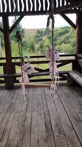 The video shows a wooden swing decorated with pink flowers and ribbons. The swing is suspended from a wooden structure, possibly a gazebo or porch, with a scenic view of rolling hills and trees in the background. The floor of the structure is made of wooden planks. The swing appears to be empty and stationary. The lighting suggests it is daytime, likely outdoors.