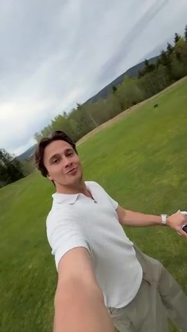 The video opens with a man holding a camera at arm's length, smiling while on a golf course. The man is wearing a white collared shirt, khaki pants, and white shoes. Behind him is a golf push cart holding a bag of clubs. Three other men are seen walking in the distance, also with golf carts. The camera is then abruptly jerked forward, blurring the scene as the man starts walking. The camera angle shifts to focus on the golf push cart, showing the hand pushing the cart forward on the green grass. The other men are now walking further ahead. The scenery includes trees and a cloudy sky.