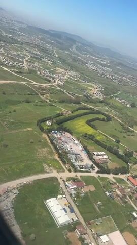 The video shows an aerial view of a landscape. The scene is dominated by green fields and scattered buildings, with a large industrial or construction site in the middle. The sky is clear and blue. In the background, there are rolling hills and a hazy cityscape. The camera appears to be moving, likely from an airplane or drone, as it provides a sweeping view of the area. Roads crisscross the landscape connecting small communities and the large site.