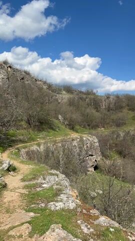 The video shows a scenic view of a canyon on a bright, sunny day. The camera pans across the landscape, revealing the rocky cliffs, the lush vegetation growing on the canyon walls, and the river flowing at the bottom of the canyon. The sky is a clear blue with scattered white clouds.