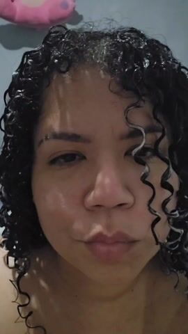 A close-up video shows a person with dark, curly, wet hair. They are looking at the camera with a slight duck face expression. The background is a gray wall, and a pink object, possibly a stuffed animal, is visible in the upper left corner. The lighting is dim.