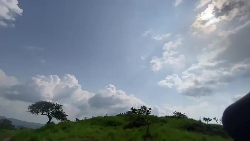 This media file showcases a scenic view from a grassy hilltop in Amaigbo, Nigeria. The primary subjects are the vast sky filled with dynamic clouds and the lush green landscape below. The sun is partially obscured by clouds, suggesting it is either early morning or late afternoon, with soft, diffused light bathing the scene. The weather appears to be fair, with fluffy white clouds scattered across a bright blue sky, interspersed with some darker, more ominous-looking clouds, hinting at the possibility of a recent or approaching shower.

The camera pans across the rolling hills, revealing patches of trees and a winding dirt path that suggests a hiking or trekking route. The terrain is uneven and covered in thick, vibrant green grass, indicating a fertile environment. In the distance, a faint hint of civilization can be seen, with scattered buildings suggesting a rural or semi-rural setting. There are no people or distinct objects visible, making the focus solely on the natural environment.

The overall mood is serene and tranquil, with a sense of vastness and natural beauty. The slight tilt of the camera and the sweeping movement across the landscape create a dynamic and immersive viewing experience. The media file appears to be a video recording, possibly a timelapse or a continuous shot of the scenery. No text is visible within the media file.