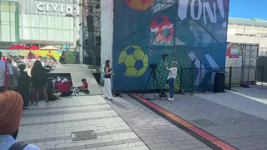 A group of people are gathered outdoors in front of a large banner with soccer balls on it. A woman with dark hair is standing holding a microphone and speaking to a woman operating a video camera on a tripod. Several children are sitting on a ramp. In the background, there are buildings and more people. The ground is paved with interlocking bricks.