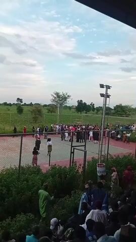 The video shows an outdoor gathering of people on what appears to be a tennis court. The court is enclosed by a fence. A crowd of people are standing around, some are walking, and a few are in the center of the court. The environment is green with trees and grass visible in the background. The sky is cloudy. There are lights on poles on the side of the court. The perspective suggests the video is taken from a slightly elevated position, possibly from a spectator area. The time of day appears to be late afternoon or early evening. The people are dressed in various types of clothing, some in what looks like traditional attire. There are no overt signs of distress or conflict.
