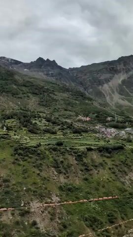 A drone flies over a mountainous landscape with terraced fields and sparse vegetation under a cloudy sky. The camera pans to the right, revealing more of the green, rocky hills and a snow-capped mountain peak in the distance. The footage appears to be taken during the daytime.