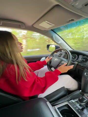 A young woman with long brown hair drives a Toyota Land Cruiser on a rural road. She wears a red sweatshirt and light-colored pants. The interior of the car is black leather. The scenery outside the window includes trees and grass. She appears to be singing along to music.