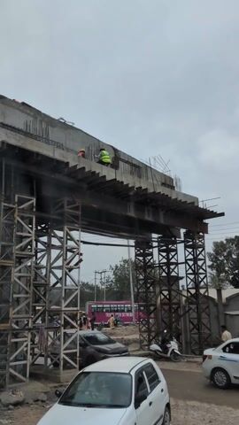 The camera captures a construction site where an overpass is being built. The overpass is supported by a complex scaffolding system made of steel beams. Two construction workers, one wearing a yellow safety vest and the other an orange hard hat, are on top of the overpass, working on the structure. Below, there is street traffic including cars, scooters, and a pink bus with "TRAVEL WITH SETHI BUS" visible. Several pedestrians can be seen walking on the side of the road, including a man in a white outfit walking to the right of the frame. The sky is overcast and gray.
