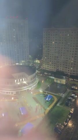 The video shows a night view of a residential area, possibly after rain, with streetlights illuminating the scene. Tall apartment buildings, one lighter colored and modern, the other darker and older, dominate the background. In the mid-ground, a circular building with a curved facade, possibly a community center or commercial space, is visible. Adjacent to it is a playground with swings, slides, and covered areas, which appear to be blue and purple tarps. A street runs through the scene, lined with parked cars. The ground appears wet, suggesting recent rainfall. Several cars are visible in a parking lot to the right of the street. The overall atmosphere is somewhat hazy and dark, with artificial lights creating reflections and a moody ambiance.