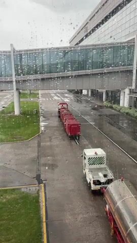 The video shows an airport tarmac on a rainy day, as seen through a window streaked with raindrops. A white baggage tug is pulling a red baggage cart train. Behind the tug is a piece of airport equipment with a conveyor belt. The tarmac is wet, and grass is visible on the left side of the frame. In the background, a modern airport terminal building with a glass walkway is visible. The sky is overcast.