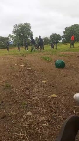 The video shows a group of people in a grassy, dirt-covered outdoor area. They appear to be wearing similar dark-colored clothing, possibly uniforms. Some individuals are holding what look like rifles. There are trees and vegetation in the background, and the sky is overcast. A green helmet is visible on the ground in the foreground. The people are spread out, and some seem to be interacting or engaged in an activity. The overall impression is that of a training exercise or practice session.