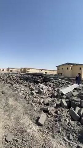 The video shows a destroyed area with piles of rubble and debris, possibly from a collapsed building or buildings. The rubble consists of grey concrete blocks and metal sheets. In the background, there are several one or two-story buildings that appear relatively intact, painted in a light tan color. Three people are walking through the rubble, presumably assessing the damage. The environment appears arid and dry, with mountains visible in the distance. The sky is clear and blue, indicating a sunny day. A white car is visible parked behind the rubble. The overall impression is one of destruction and devastation.