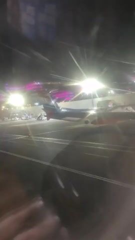 The video shows an airplane on the tarmac at night. The scene is viewed through a window, possibly from inside another airplane or a terminal building. Bright lights illuminate the tarmac, creating glare on the window. The tail of the airplane, which is blue and has a red stripe, is visible. The rest of the airplane is also visible, and it appears to be a passenger jet. There are also some lights in the background that appear to be from a terminal building or a cityscape. The audio consists of spoken instructions about emergency exits.