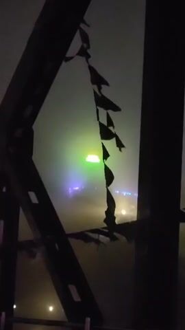 The video shows a close-up of dark, metal structures, possibly part of a bridge or building, in the foreground. Partially obscured by these structures, in the background, is a hazy, dimly lit scene. A bright green light emanates from a central point, diffused by the fog or mist. Below this green light, there are hints of other colorful lights, primarily blue and purple, and some smaller, scattered white lights that might be distant lights or lights on the ground. A string of torn, dark fabric or flags hangs from the metal structure in the foreground, partially obscuring the background. The overall atmosphere is dark, moody, and somewhat eerie due to the low visibility and the strange lighting. The fog or mist is a significant element, softening the lights and creating a diffused, almost otherworldly effect.