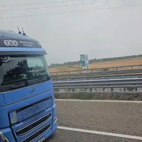 The video shows a blue Volvo truck driving on a highway. The truck has the logo T&A Transport on the side. The driver's side door is visible, and the interior of the cab can be seen through the windows. The truck is driving on a paved road with a guardrail on the right side. In the background, there are fields and trees. The sky is overcast. The video appears to be recorded from the perspective of someone in another vehicle driving alongside the truck.